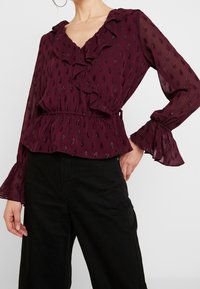 Blusa bordeaux in tessuto leggero, presenta un colletto e polsini arricciati, con un sottile motivo a foglia metallizzata e dettaglio in vita svasato.