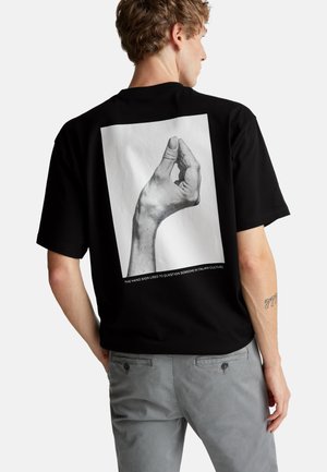 Camiseta de algodón negra con un gran gráfico blanco de una mano con un diseño intrincado. Mangas cortas y corte relajado. Pantalones grises.