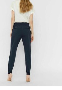 Marinblå avsmalnande byxor med elastisk midja, sidofickor och en mjuk textur, parat med nude högklackade skor och en avslappnad vit skjorta.