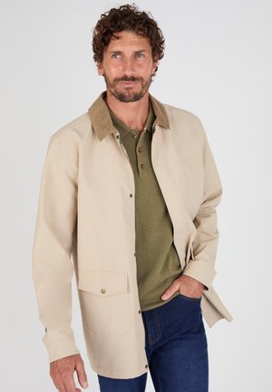 Giacca impermeabile beige con colletto in velluto a coste, tasche anteriori e chiusure a scatto, indossata sopra una polo verde e jeans scuri.