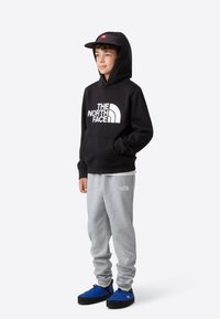 Črna jopa s kapuco z belim logotipom, sive športne hlače in modra obutev. Kodrasti lasje, stoječi položaj, minimalistično ozadje.