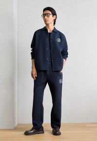 Άνδρας που φοράει σκούρο μπλε μπουφάν με κουμπιά και ταιριαστό παντελόνι με λογότυπα, μαύρα γυαλιά με σκελετό και καφέ μοκασίνια, στέκεται μπροστά σε απλό τοίχο.