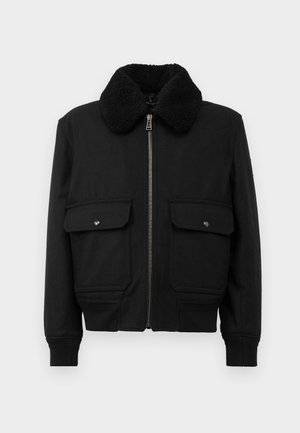 Black jacket with a textured black shearling collar, front zipper, and two chest pockets featuring snap closures. Smooth fabric, fitted design.