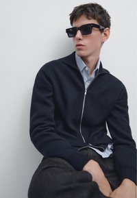 Mörk marinblå zip-up-tröja i mjukt material, lager ovanpå en ljusblå randig skjorta. Svarta rektangulära solglasögon ger kontrast.