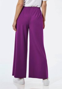 Un pantalon large violet en tissu lisse et léger. Il met en avant une taille haute et une silhouette ample et fluide. Porté avec des chaussures blanches.