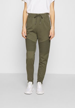 Personne portant un pantalon côtelé vert olive aux genoux avec taille à cordon et baskets blanches, debout devant un fond gris uni.