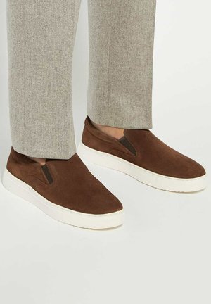 Brown suede slip-on sneakers with elastic side panels and white rubber soles, paired with light grey tailored pants.