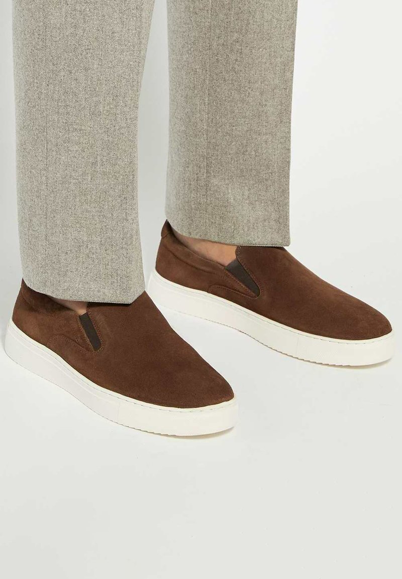 Brown suede slip-on sneakers with elastic side panels and white rubber soles, paired with light grey tailored pants.