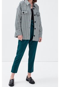 Zwart-witte houndstooth jas met grote knopen, gecombineerd met teal taps toelopende broek en zwarte loafers met metalen studs.