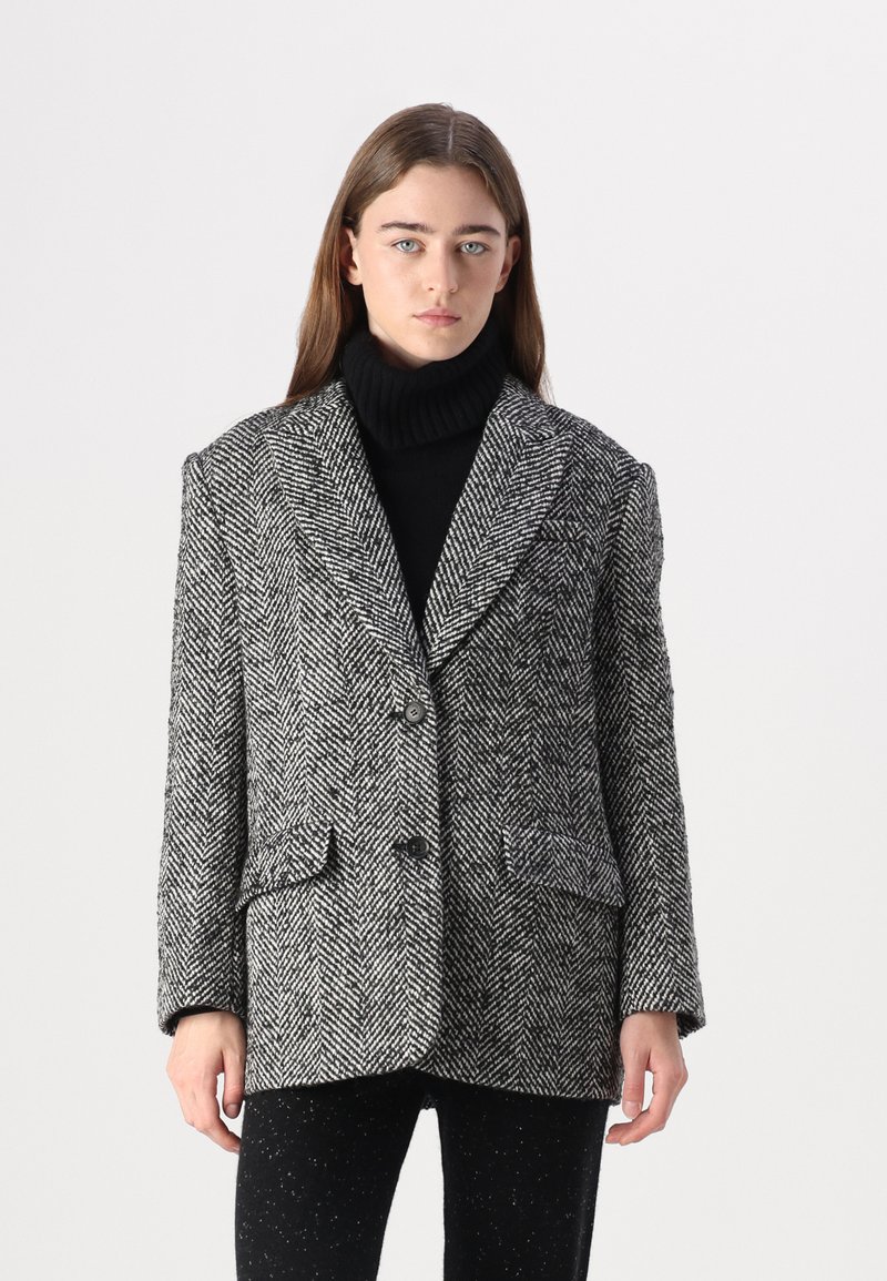 Blazer à motif chevron noir et blanc avec col à revers, boutons et deux poches avant, porté sur un col roulé noir.