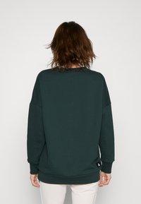 Felpa verde scuro con una vestibilità rilassata, spalle scese e polsini a coste. Un piccolo etichetta nera con logo è visibile vicino all'orlo.