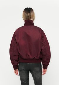Burgundy bomber jacket with a high collar, ribbed cuffs, and hem; smooth texture; features padded design and tonal branding on back.