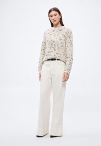 Gestrickter Pullover in Creme mit strukturiertem Muster; kombiniert mit weit geschnittenen hellen Hosen und schwarzen Absatzschuhen. Dezenter Gürtelakzent.