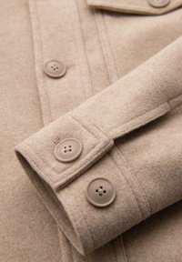 Sezione di un cappotto beige in lana con un primo piano del polsino, che presenta due bottoni rotondi e una finitura morbida e testurizzata.
