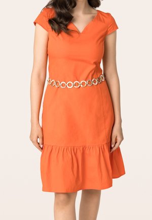 Kobieta w pomarańczowej sukience do kolan z krótkimi rękawami i ozdobnym złotym paskiem łańcuchowym w kształcie koła w talii.