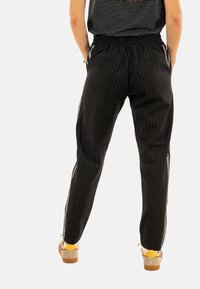 Zwarte pantalon met streepdessin, een elastische tailleband, zijzakken en witte zijkanten; model draagt gele en crèmekleurige klompen.