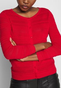 Femme les bras croisés portant un cardigan rouge vif texturé à boutons et un pantalon en cuir noir, sur un fond uni.