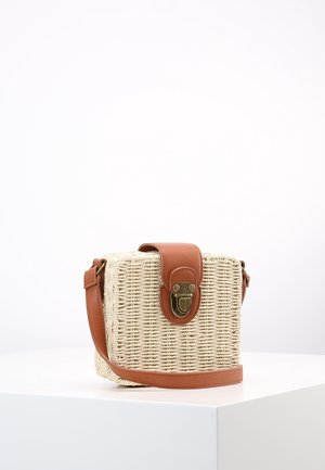 Quadratisch gewebte Strohtasche mit braunem Lederriemen und Klappe, versehen mit einem Messingverschluss, auf einer weißen Fläche vor schlichtem Hintergrund liegend.