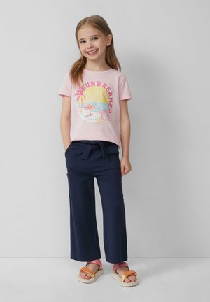 Lichtroze t-shirt met een kleurraf beach graphic, gecombineerd met donkerblauwe wijde broeken. Heeft oranje, roze en witte sandalen.