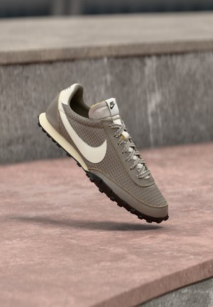 Pantof sport Nike bej și alb cu partea superioară din plasă și talpă neagră texturată, poziționat pe călcâi pe o suprafață de piatră roz.