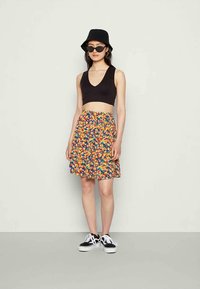 Mulher usando um chapéu preto tipo bucket, top cropped preto sem mangas, saia floral colorida, óculos de sol pretos e ténis preto e branco, de pé contra uma parede branca.