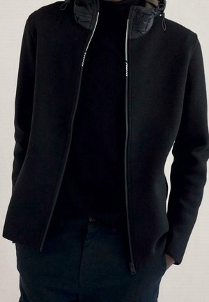 Mann trägt eine schwarze Zip-up-Jacke über einem schwarzen Hemd, die Hände in den Taschen, steht vor einem einfarbigen hellen Hintergrund.