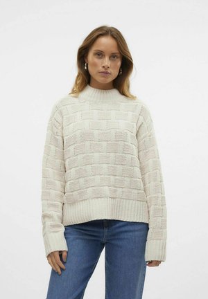 Pull en tricot crème avec un motif texturé en tissage de panier, doté d'un col montant et de poignets côtelés, associé à un jean en denim bleu.