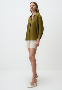 Blusa verde oliva a maniche lunghe con colletto, abbinata a pantaloni corti beige chiaro e scarpe con tacco bianche. Tessuto liscio.