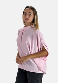 Blusa over-size rosa con collo alto, texture morbida, maniche corte e pieghe sottili. Abbinata a jeans grigio scuro.