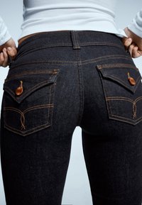 Ciemne dżinsy z denimu mają dopasowany krój z pomarańczowymi przeszyciami i dwoma tylnymi kieszeniami. Każda kieszeń jest zapinana na guzik i ozdobiona wzorem dekoracyjnym.