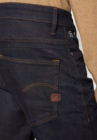 Vaqueros de mezclilla oscura con costuras naranjas, que cuentan con una etiqueta de cuero, bolsillos estándar y un diseño ajustado. La vista trasera muestra detalles.