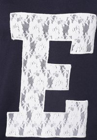 Tessuto blu navy con un grande disegno in pizzo bianco a forma di lettera "E", caratterizzato da motivi floreali sul pizzo e una finitura testurizzata.