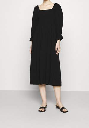 Femme portant une longue robe noire avec des manches bouffantes et un décolleté carré, associée à des sandales noires à enfiler, debout sur un sol gris.