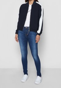 Chaqueta bomber de la marina con rayas blancas, cierre de cremallera en la parte delantera, puños de canalé. Combinada con jeans ajustados azules y zapatillas blancas. Atuendo clásico y casual.