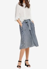 Blouse blanche à coupe ample avec détails en dentelle ; jupe midi rayée bleu et blanc avec ceinture à noeud et poches latérales ; assortie à des escarpins noirs.