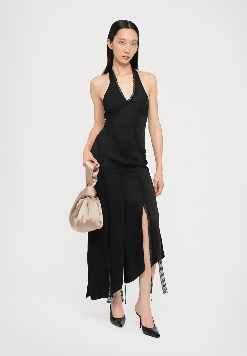 Black halter neck dress with lace detailing, fitted silhouette, side slit, and contrast stitching. Paired with a beige satin bag and black heels.