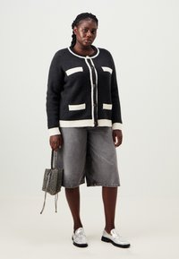 Μαύρος και κρεμ cardigan με κλείσιμο κουμπιού, που διαθέτει δύο μπροστινές τσέπες. Γκρι τζην σορτσάκια και γυαλιστερά ασημί μοκασίνια. Μικρή τσάντα με σχέδιο.