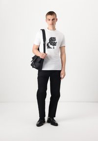Hvit T-skjorte med svart grafisk design, kombinert med svarte bukser og svarte sko. Modellen holder en svart crossover-veske.