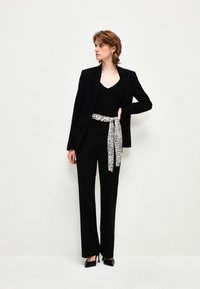 Zwarte op maat gemaakte blazer over een zwart topje, gecombineerd met zwarte broeken. Een wit-zwarte gestructureerde ceintuur accentueert de taille. Zwarte hakken maken de look compleet.
