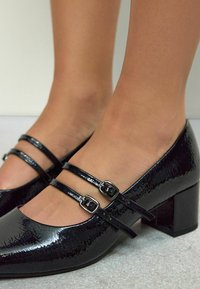 Black patent leather Mary Jane heels with two adjustable straps, square toe, block heel, and a textured surface.