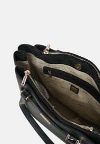 Borsa nera con esterno testurizzato, dotata di una chiusura con zip e molteplici scomparti. Interno foderato in beige con logo "GUESS" ripetuto.