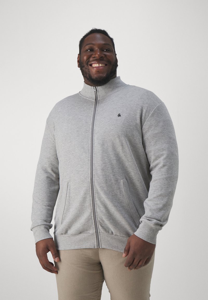 Sudadera gris con cremallera y cuello alto, mangas largas, bolsillos frontales y un pequeño logotipo en el pecho. Tejido suave con puños acanalados.