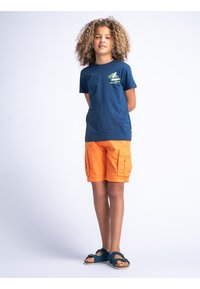 Marineblauw katoenen T-shirt met een groene grafische opdruk op de borst, gecombineerd met oranje cargoshorts en teal sandalen met een gespsluiting.