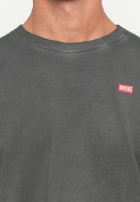 Gros plan sur une personne portant un t-shirt à col rond gris foncé avec un petit logo Diesel rouge et blanc sur le côté supérieur gauche de la poitrine.