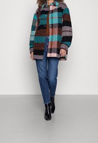 Yksivärinen villasekoiteshirt-jakku, jossa on monivärinen ruutukuvio turkoosin, ruskean, mustan ja vaaleanpunaisen sävyissä. Varustettu nappilukolla ja kauluksella.