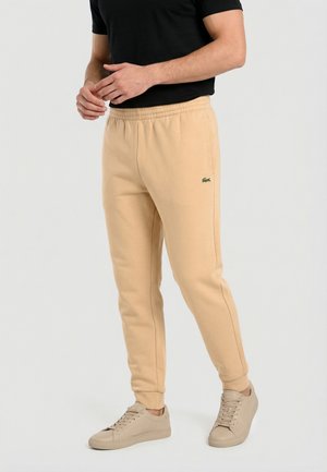 Homme portant un pantalon de survêtement beige avec le logo Lacoste, des baskets beiges et un t-shirt noir à manches courtes, debout devant un fond clair uni.