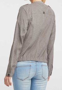Chaqueta gris, ligera, con una cintura ajustada, botón decorativo en la parte posterior y mangas largas. Llevada con jeans de color azul claro.