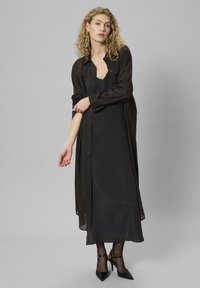 Donna con capelli biondi ricci che indossa un lungo abito nero, una camicia nera trasparente con bottoni, collant neri e tacchi alti neri, in piedi contro uno sfondo grigio.