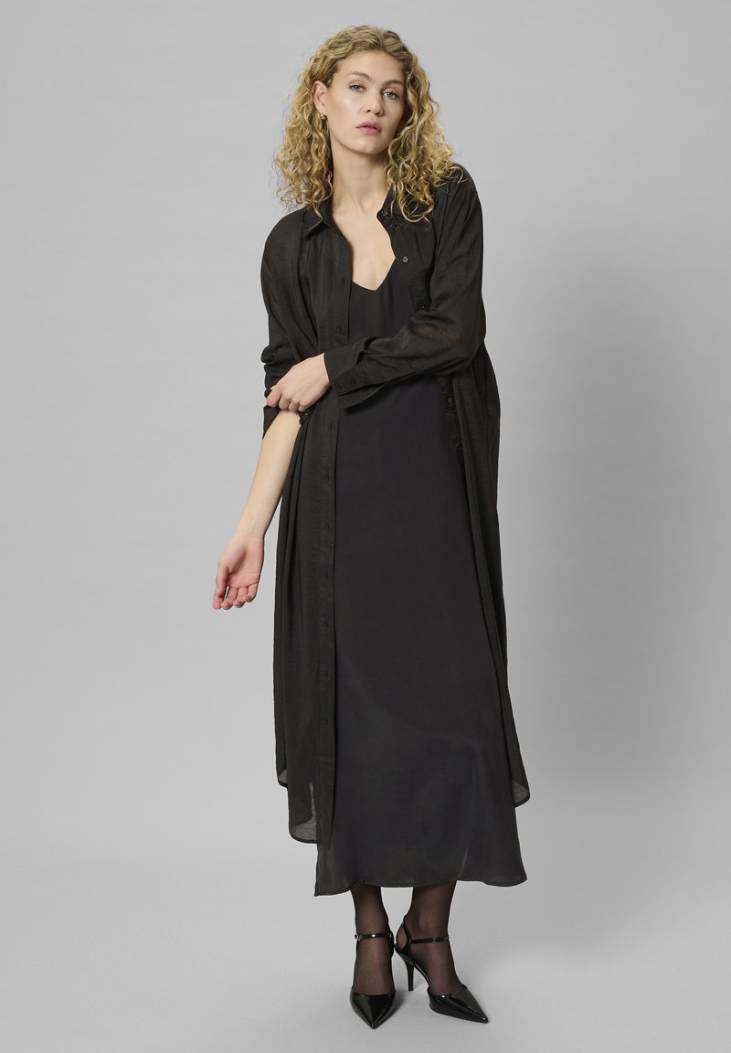 Femeie cu păr blond creț, îmbrăcată într-o rochie lungă neagră, cămașă transparentă neagră cu butoni, colanți negri și pantofi cu toc negri, stând în fața unui fundal gri.