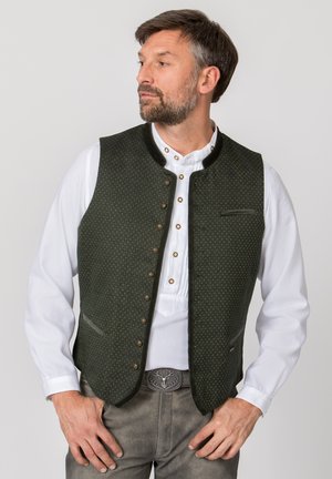 Mann trägt eine dunkelgrüne traditionelle Weste über einem weißen Hemd mit dekorativen Knöpfen und graue Hose mit Hirsch-gravierter Gürtelschnalle.
